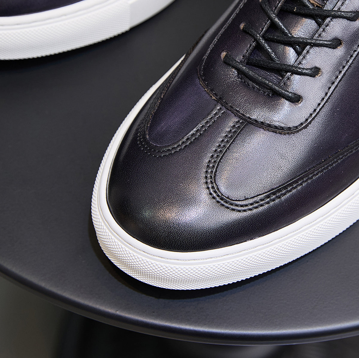 Close-up of dark leather sneaker with black laces, stitched panels, and white sole.