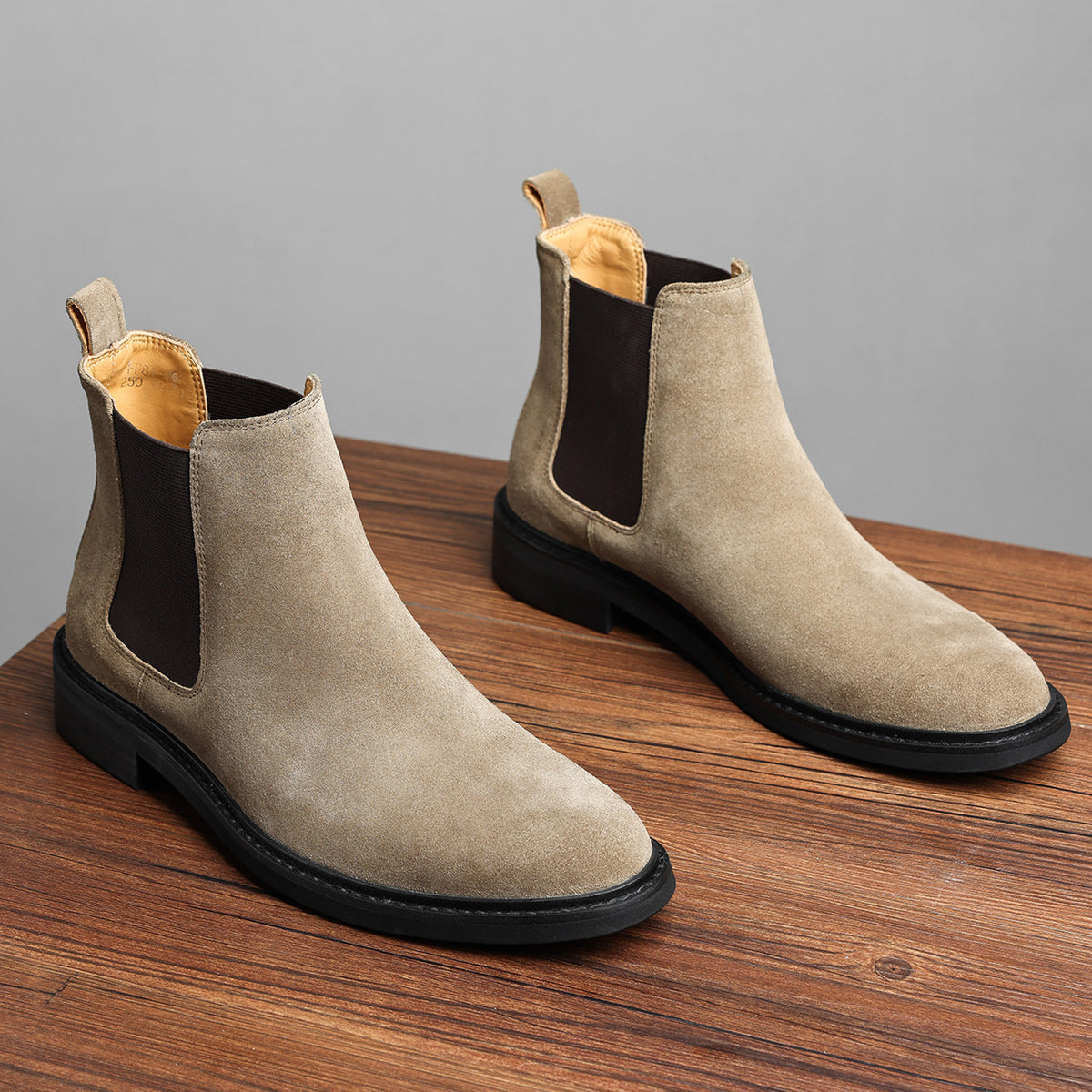 Taupe Chelsea dress boots with side elastic gore and low stacked heel on wood surface.