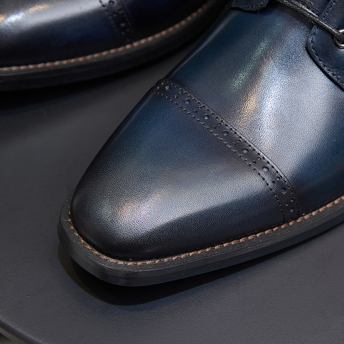 Leather boot toe close-up with brogue perforations and tan welt, dark blue.