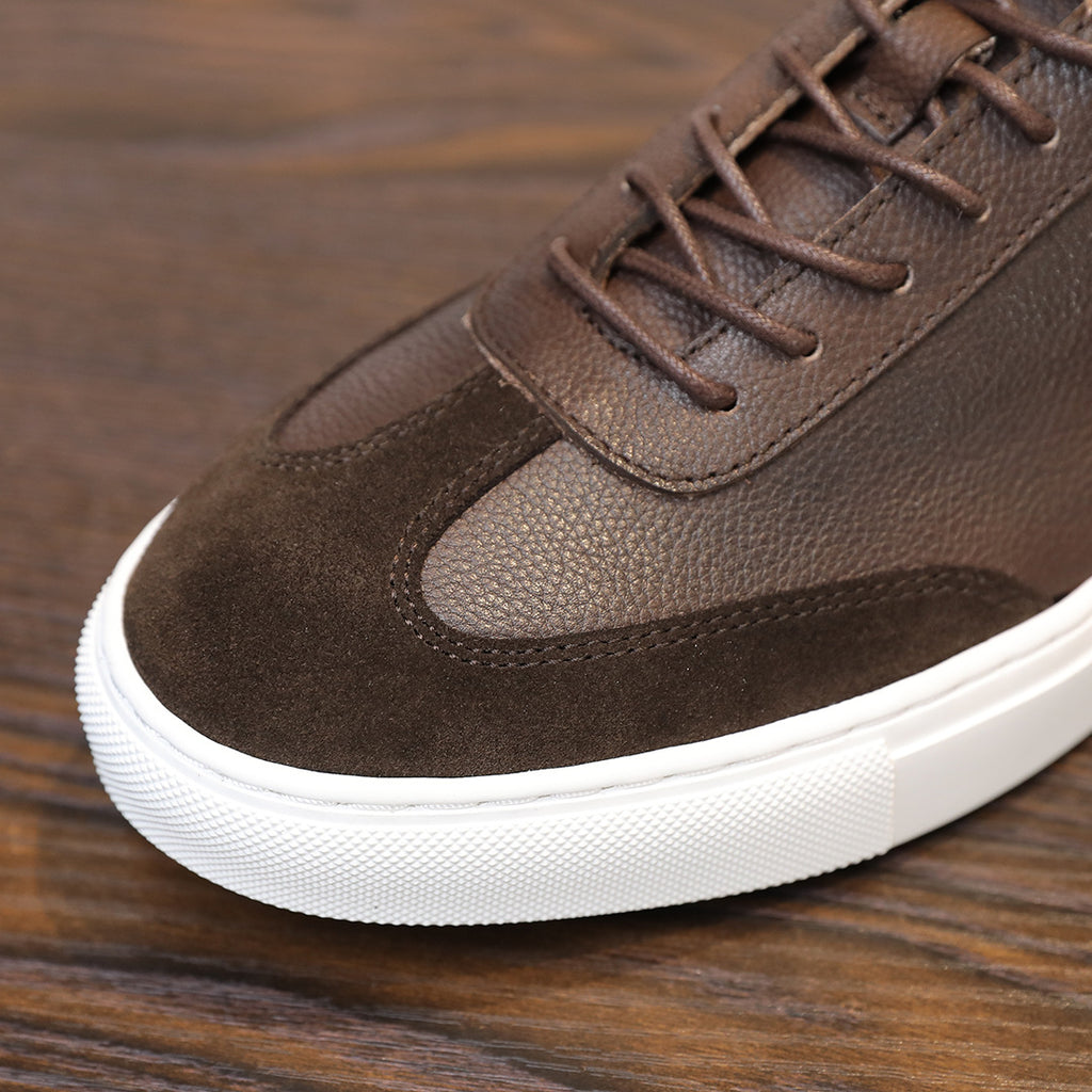 Men's dark brown leather sneaker close-up with laces, suede toe panel, and white sole.