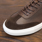 Men's dark brown leather sneaker close-up with laces, suede toe panel, and white sole.