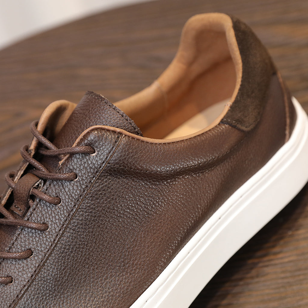 Men's dark brown leather sneaker close-up with tan lining, laces, and white sole.