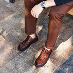Brown lace-up leather sneakers with perforations and chunky sole, worn with trousers.