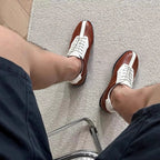 Brown leather sneakers with white laces and side panels, worn with dark trousers.